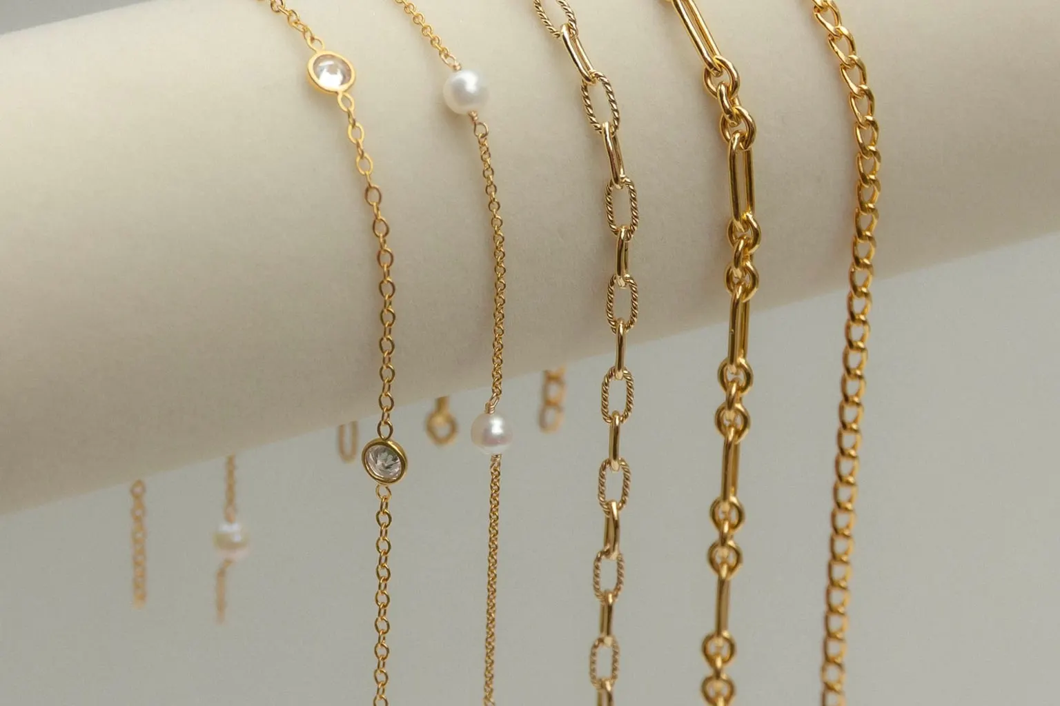 Five gold chain styles draped over a cream jewelry bolster, from crystal and pearl stations to paperclip and cable links