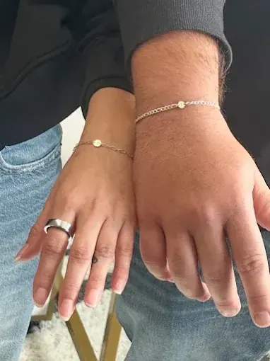 Two people standing side by side in denim, each wearing a matching thin silver chain bracelet with a small disc