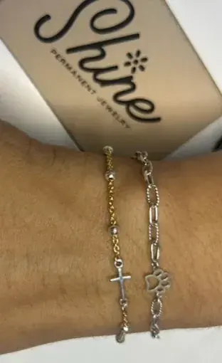 Two bracelets on a wrist, one gold with a small cross charm and one silver with a paw-print charm, Shine card behind