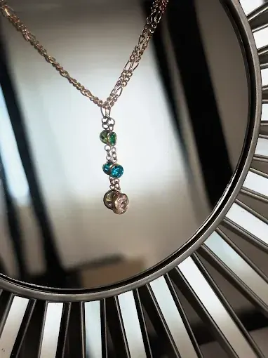Gold necklace with a three-stone drop pendant in green, turquoise, and clear, hanging in front of a sunburst mirror