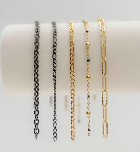 Five jewelry chains hanging vertically from a white bar, including dark links and gold Figaro, beaded, and paperclip styles
