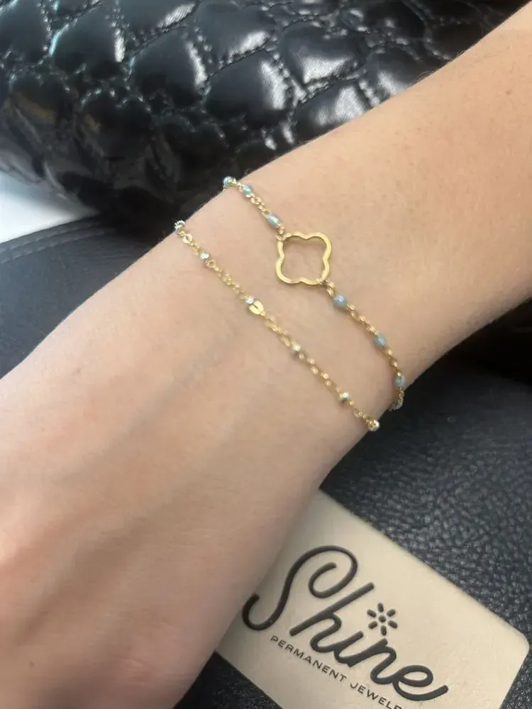 Two gold bracelets on a wrist, one with a clover charm and pale beads, Shine Permanent Jewelry card nearby