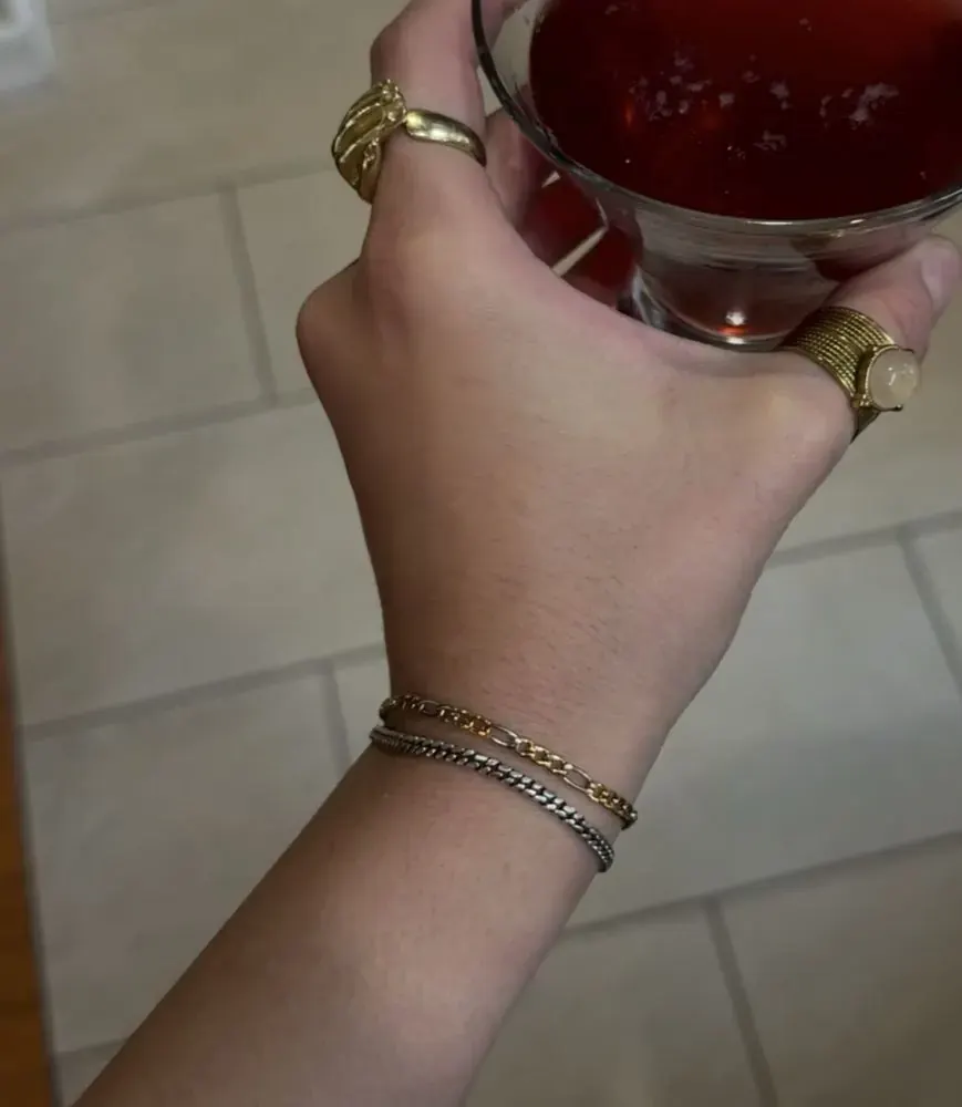 Hand holding a glass of red wine, wearing stacked gold and silver chain bracelets and two gold rings