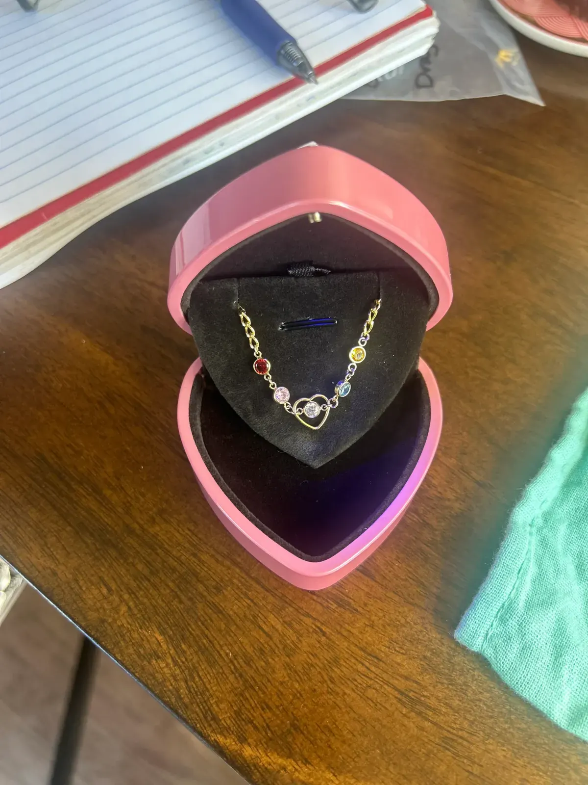 Gold necklace with colorful bezel-set stones and an open-heart charm inside a pink heart-shaped jewelry box