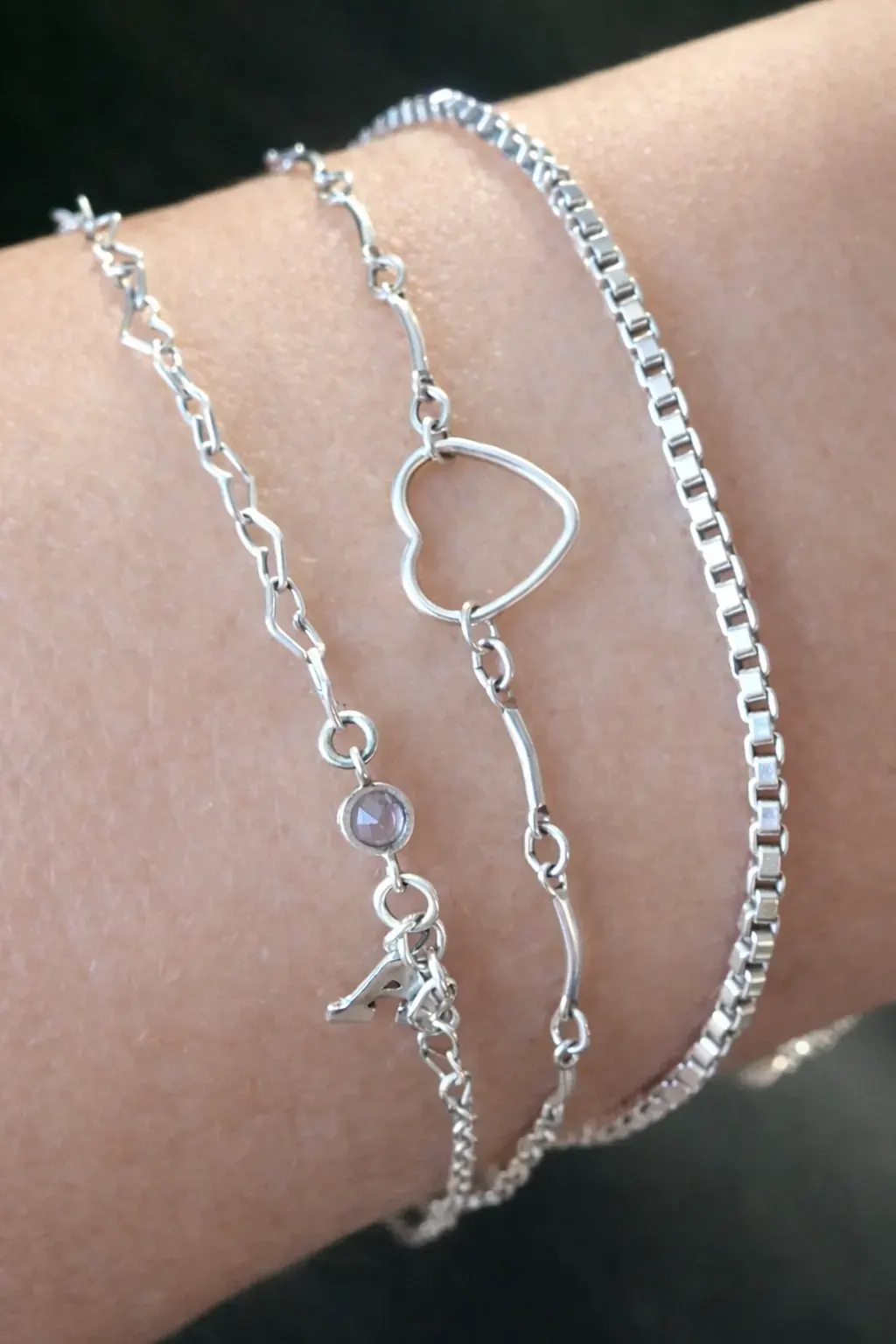 Three layered silver bracelets on a wrist: an initial A with a purple stone, an open-heart bar chain, and a box chain