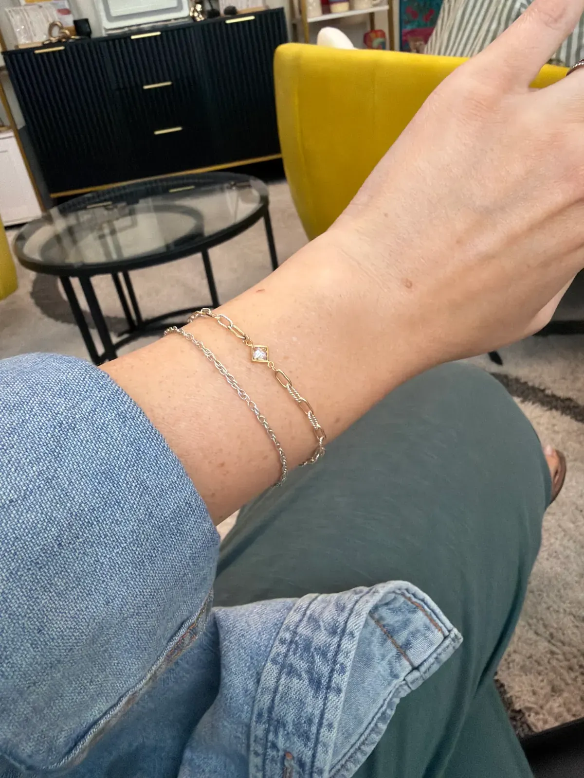 Wrist in a denim sleeve wearing a gold paperclip bracelet with a small stone and a thin silver rope chain