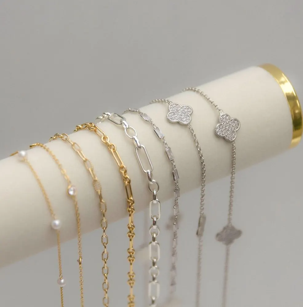 Eight gold-filled and sterling silver bracelets draped on a cream jewelry bolster, from pearl and paperclip chains to clover charms.