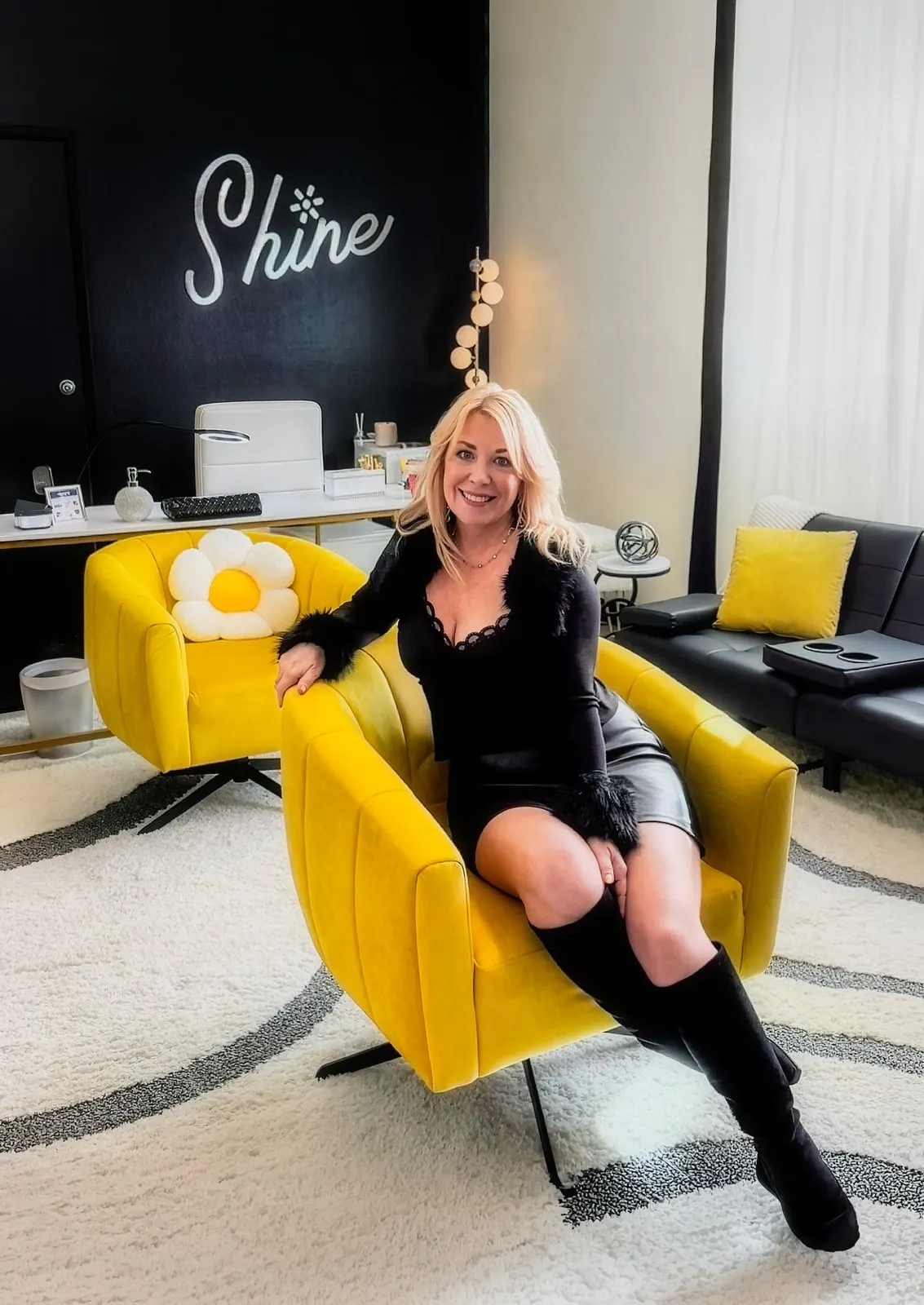 Lynette Page, owner of Shine Permanent Jewelry, seated in the studio lounge in front of the Shine script on the wall.